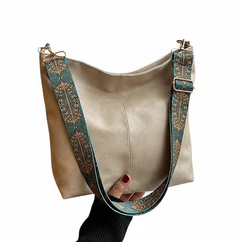 Beige handbag with decorative strap held by a person in front of a window.