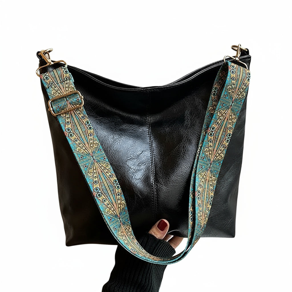 Black leather handbag with a decorative blue strap held by a person in front of a window.